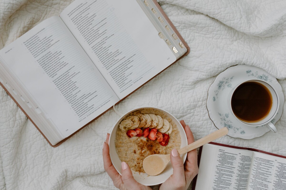 a person holding a bowl of cereal beside the open book and coffee